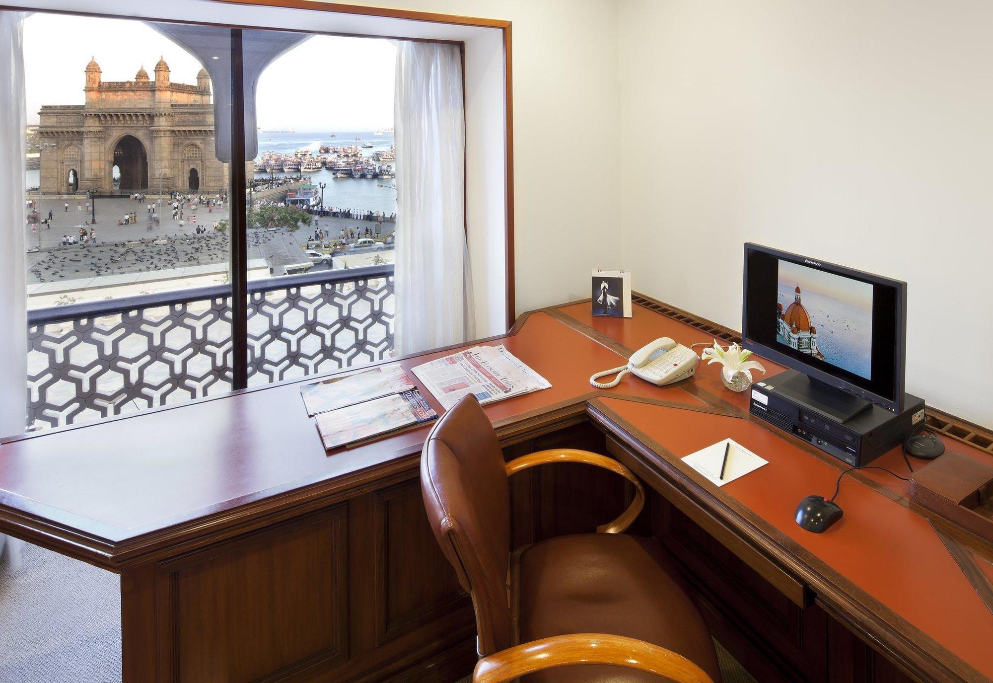 The Taj Mahal Tower, Mumbai Hotel Kültér fotó The photo shows a workspace with a wooden desk and an ergonomic chair. On the desk, there is a computer monitor, a telephone, and some stationery items. A window provides a view of an outdoor scene, possibly featuring water and structures or building